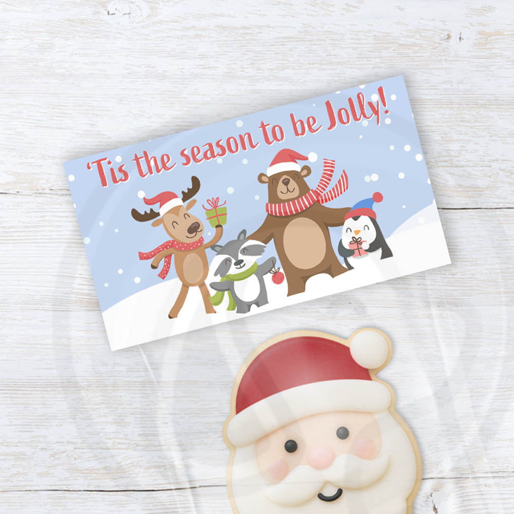 Printable “’Tis the Season to Be Jolly” holiday treat topper featuring reindeer, bear, penguin, and badger in scarves and Santa hats on a snowy blue background — perfect for folding over 4" and 6.5" holiday treat bags.