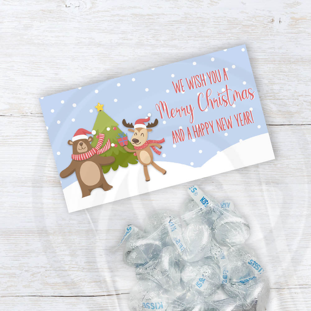Printable Christmas treat bag topper featuring a cute reindeer and bear in scarves dancing by a Christmas tree with “We wish you a Merry Christmas and a Happy New Year” text on a snowy blue background.
